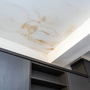 Large brown water stains and moisture damage spreading across a white ceiling above dark wooden cabinets.