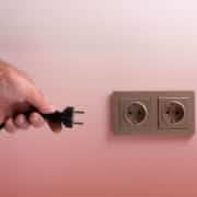Close-up of a hand about to plug a black electrical cord into a wall outlet against a light pink background.