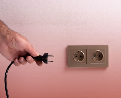 Close-up of a hand about to plug a black electrical cord into a wall outlet against a light pink background.