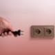 Close-up of a hand about to plug a black electrical cord into a wall outlet against a light pink background.