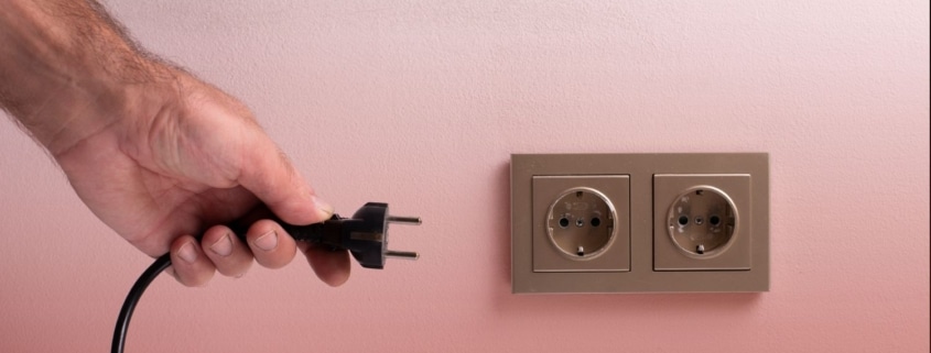 Close-up of a hand about to plug a black electrical cord into a wall outlet against a light pink background.