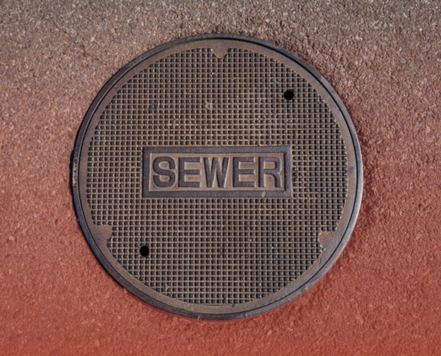Overhead view of a round metal manhole cover stamped with the word "SEWER" set in asphalt pavement.