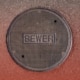 Overhead view of a round metal manhole cover stamped with the word "SEWER" set in asphalt pavement.
