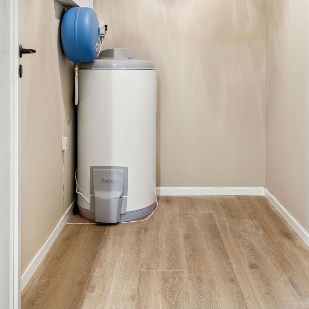 A white residential water heater with a blue expansion tank and supply lines installed in a utility closet.