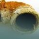 Close-up cross-section of a deteriorated Orangeburg sewer pipe showing delaminating, brittle layers and heavy rust and mineral buildup around the opening.